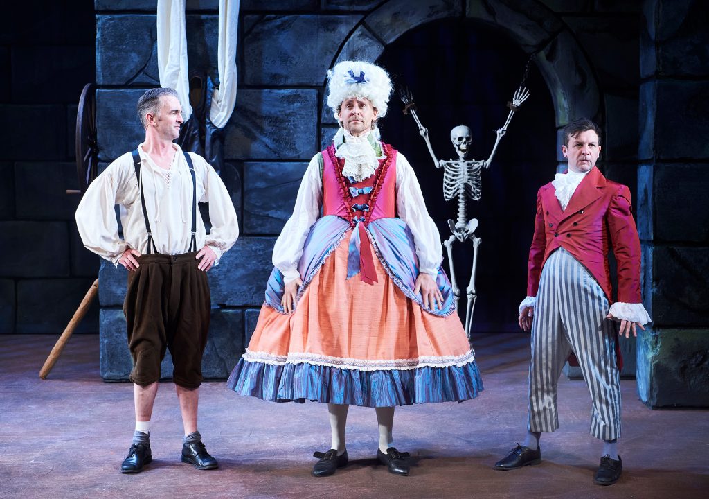 Production photo from The Play What I Wrote of the two main men with the guest star Tom Hiddleston is in a dress and a large wig. They're in a cellar with a fake skeleton in the background.