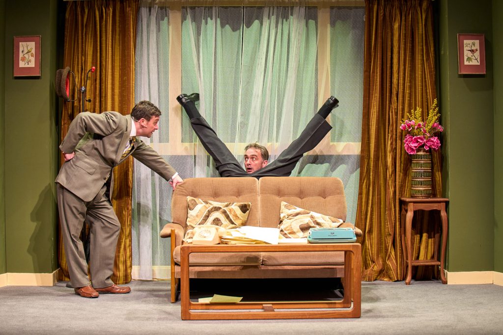 Production photo from The Play What I Wrote of two men, one is behind a sofa with his legs behind his head, the other is looking at him