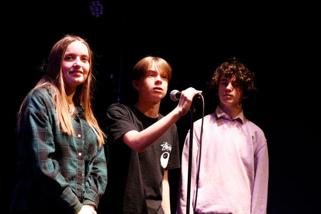 Production photo of two boys and one girl around a microphone.