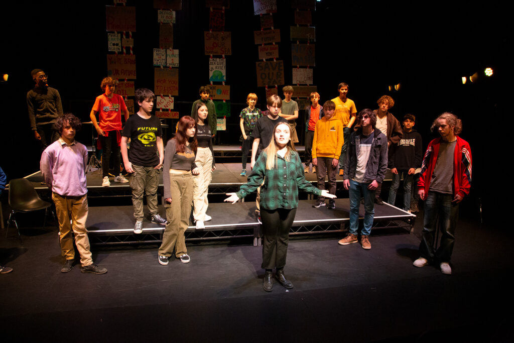 Production photo of a group of teenagers standing facing the audience on a stage. They are in a semi-circle formation around a girl who has her arms raised slighty.